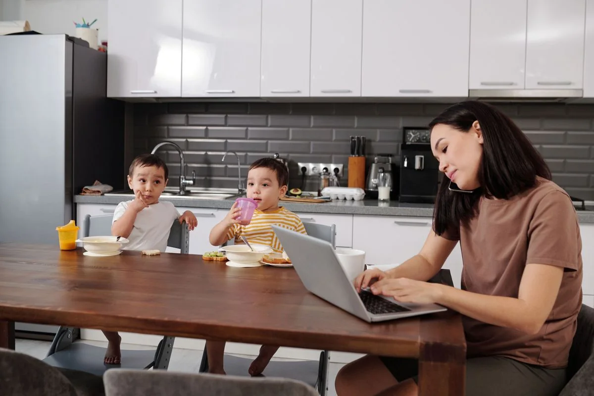 Dos niños pequeños desayunando en la mesa de la cocina mientras su madre trabaja en la laptop