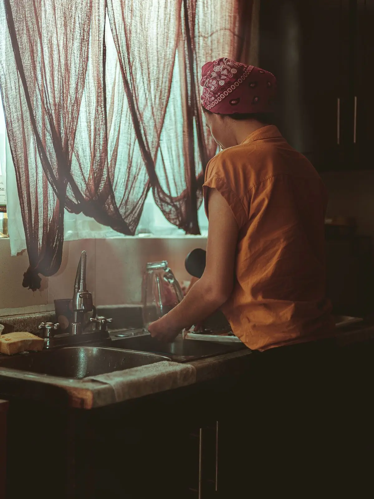 Mujer de espaldas lavando los platos frente a una ventana con cortinas, en una cocina en penumbra