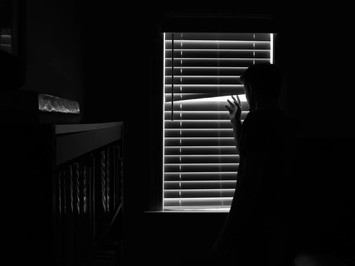 Silueta de una persona que separa las persianas para mirar afuera, en una habitación a oscuras