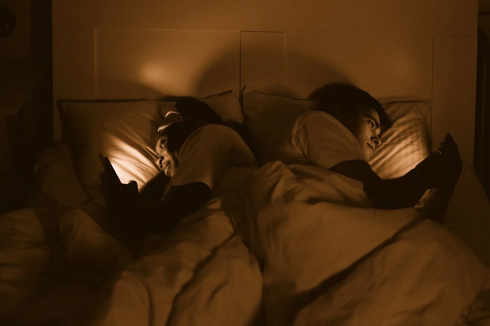 Pareja acostada en la misma cama, de espaldas, cada uno mirando su teléfono de noche