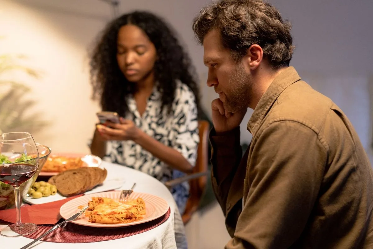 Pareja en una cena en casa: ella mira su teléfono y él, pensativo, mira hacia otro lado
