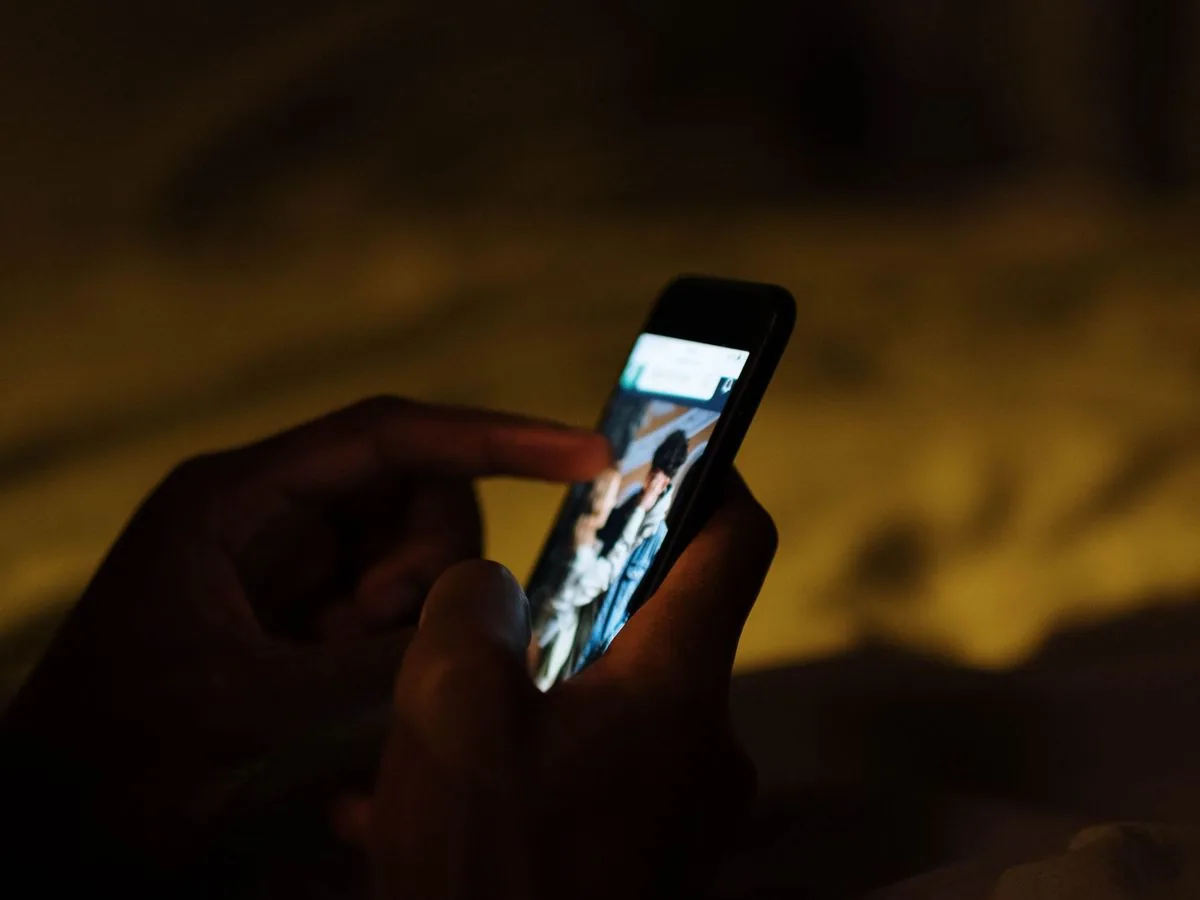 Manos que sostienen un celular encendido en la oscuridad, con un resplandor cálido de fondo