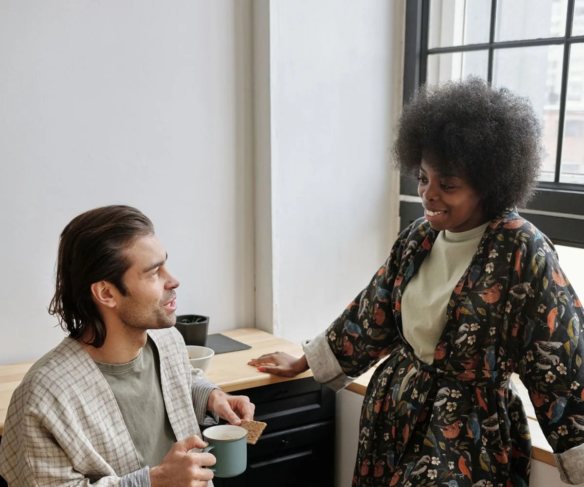 Pareja conversando en la cocina por la mañana: él habla con una taza en la mano y ella lo escucha sonriendo