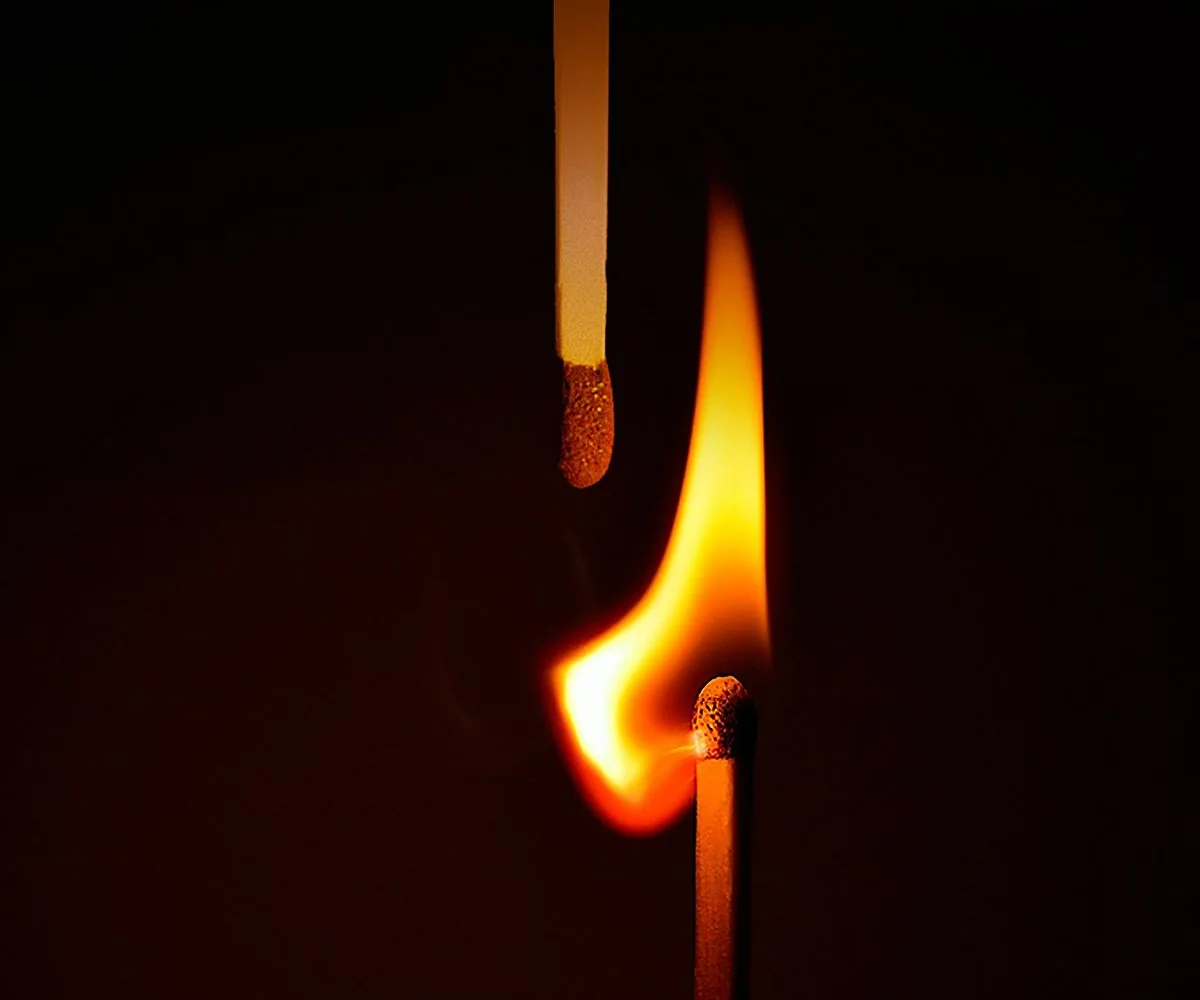 Un cerillo encendido a punto de prender otro cerillo, sobre fondo oscuro