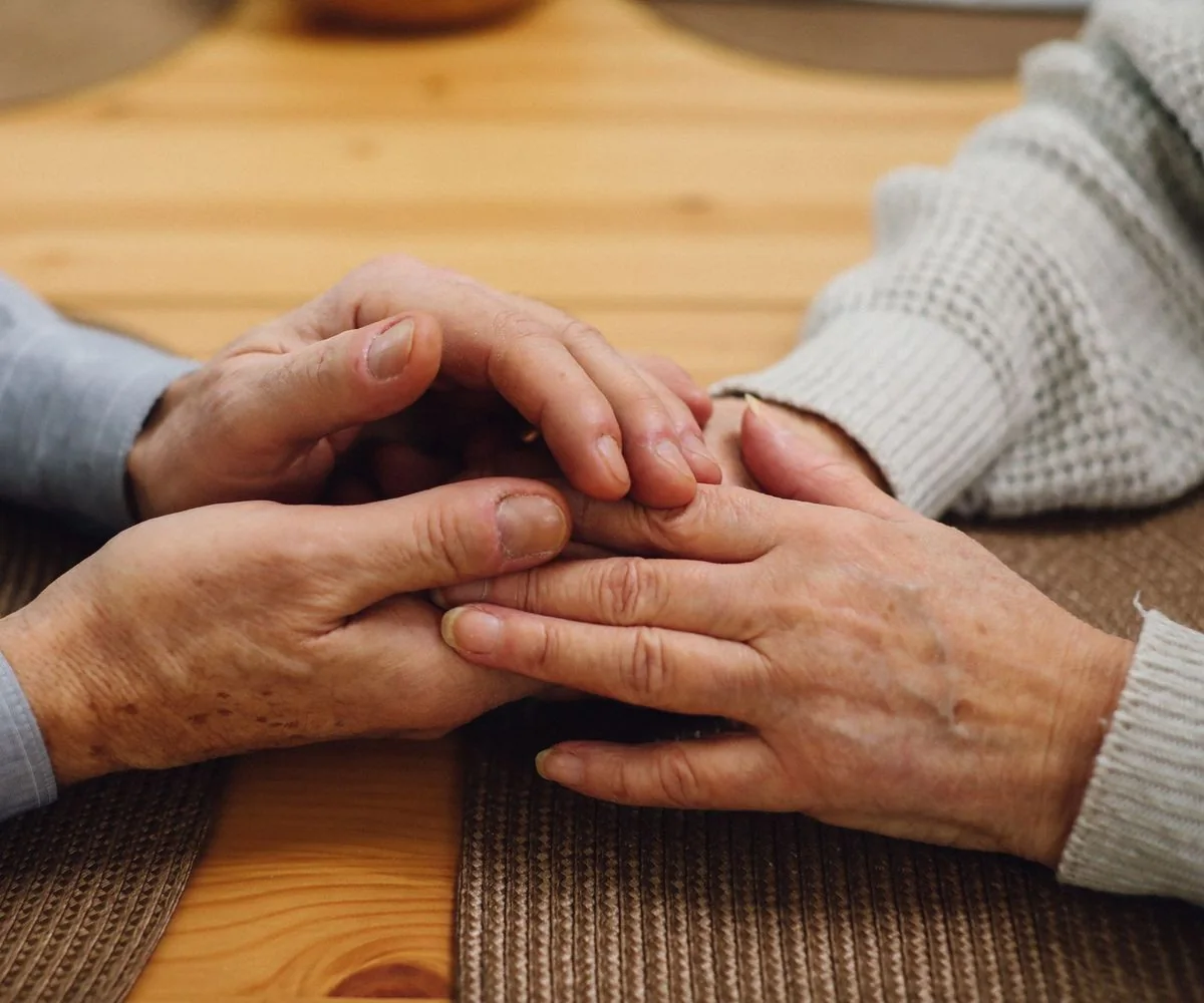 Manos de una pareja mayor entrelazadas sobre una mesa de madera, en un gesto de cuidado