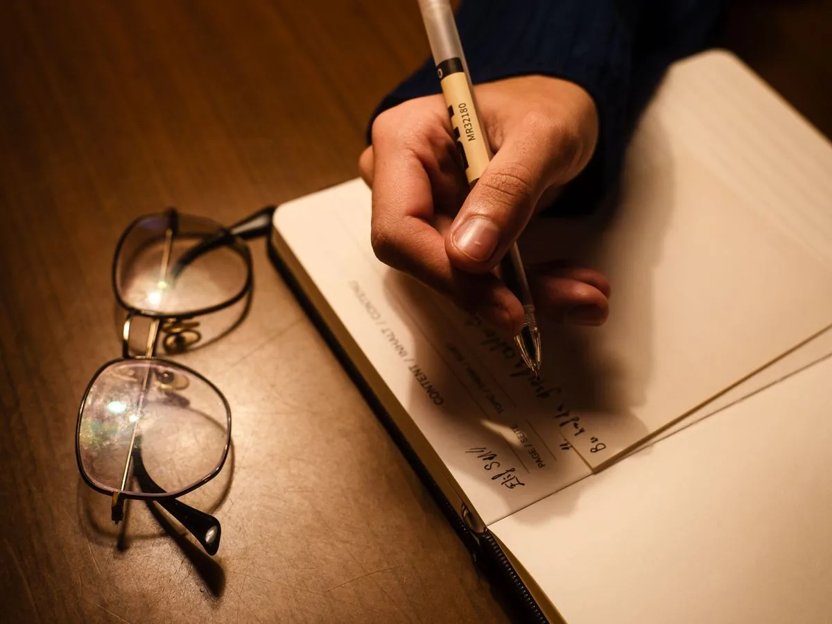 Mano que escribe en un cuaderno junto a unos lentes, bajo la luz cálida de una lámpara