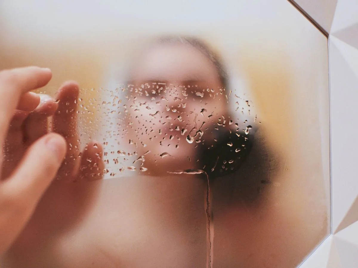 Mano que limpia un espejo empañado; detrás de las gotas, un reflejo borroso