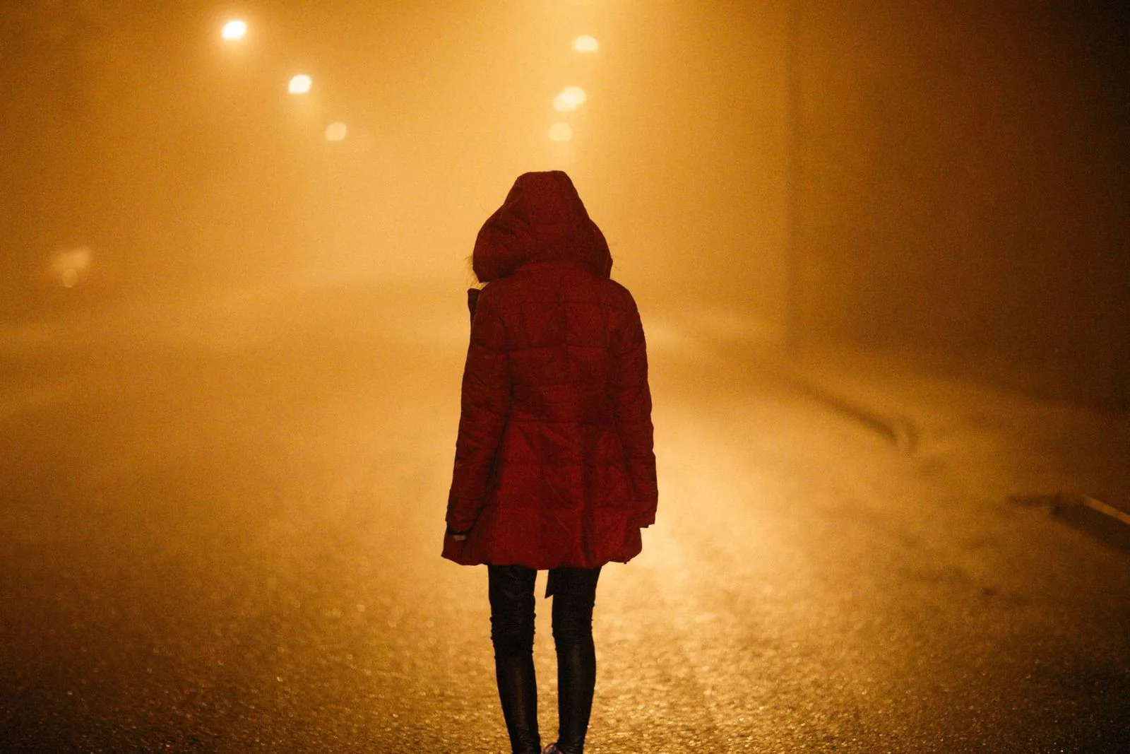 Persona de espaldas, con abrigo rojo, en una calle cubierta de niebla bajo faroles ámbar