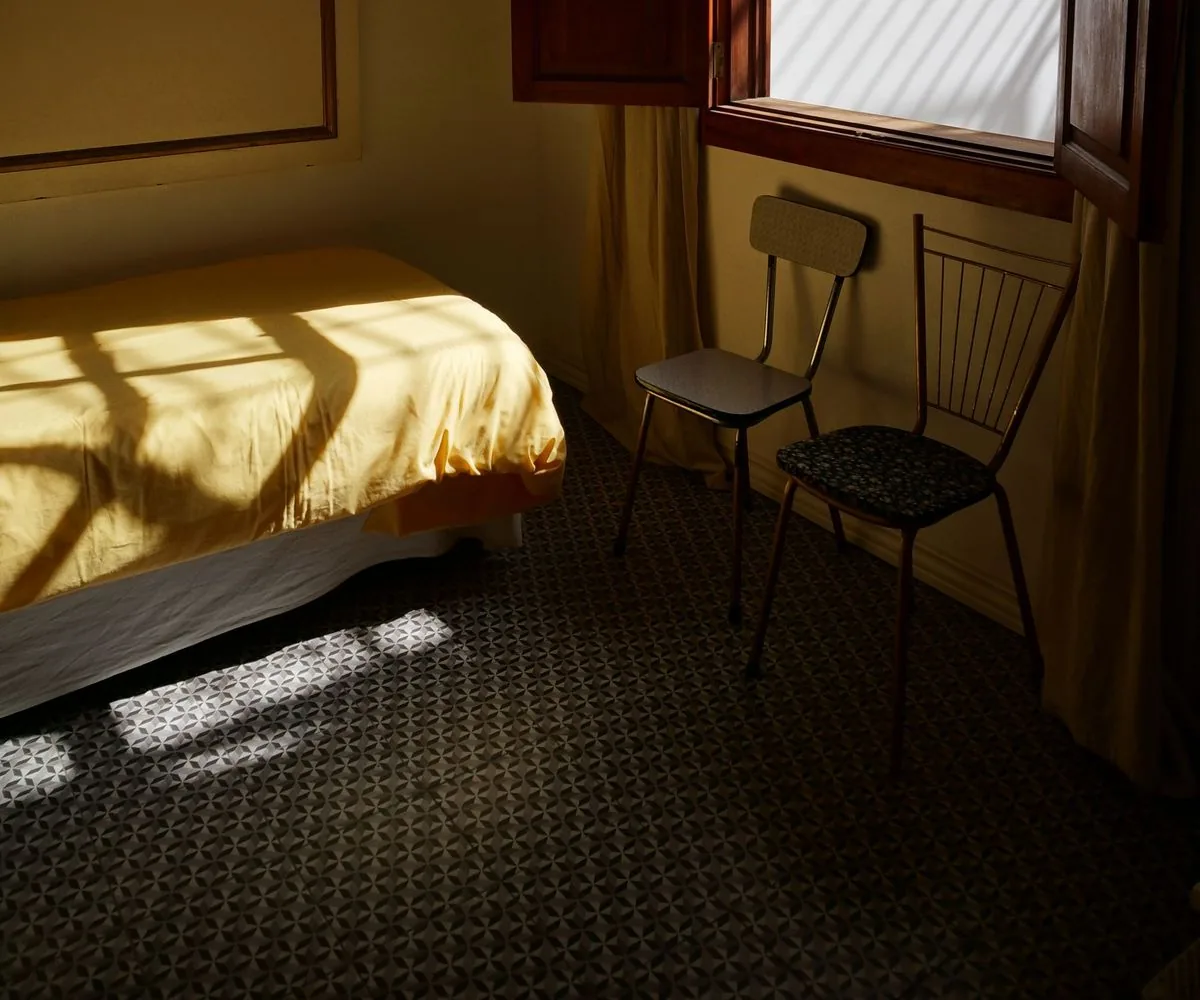 Habitación vacía con la cama tendida y dos sillas junto a la ventana, iluminada por el sol de la tarde
