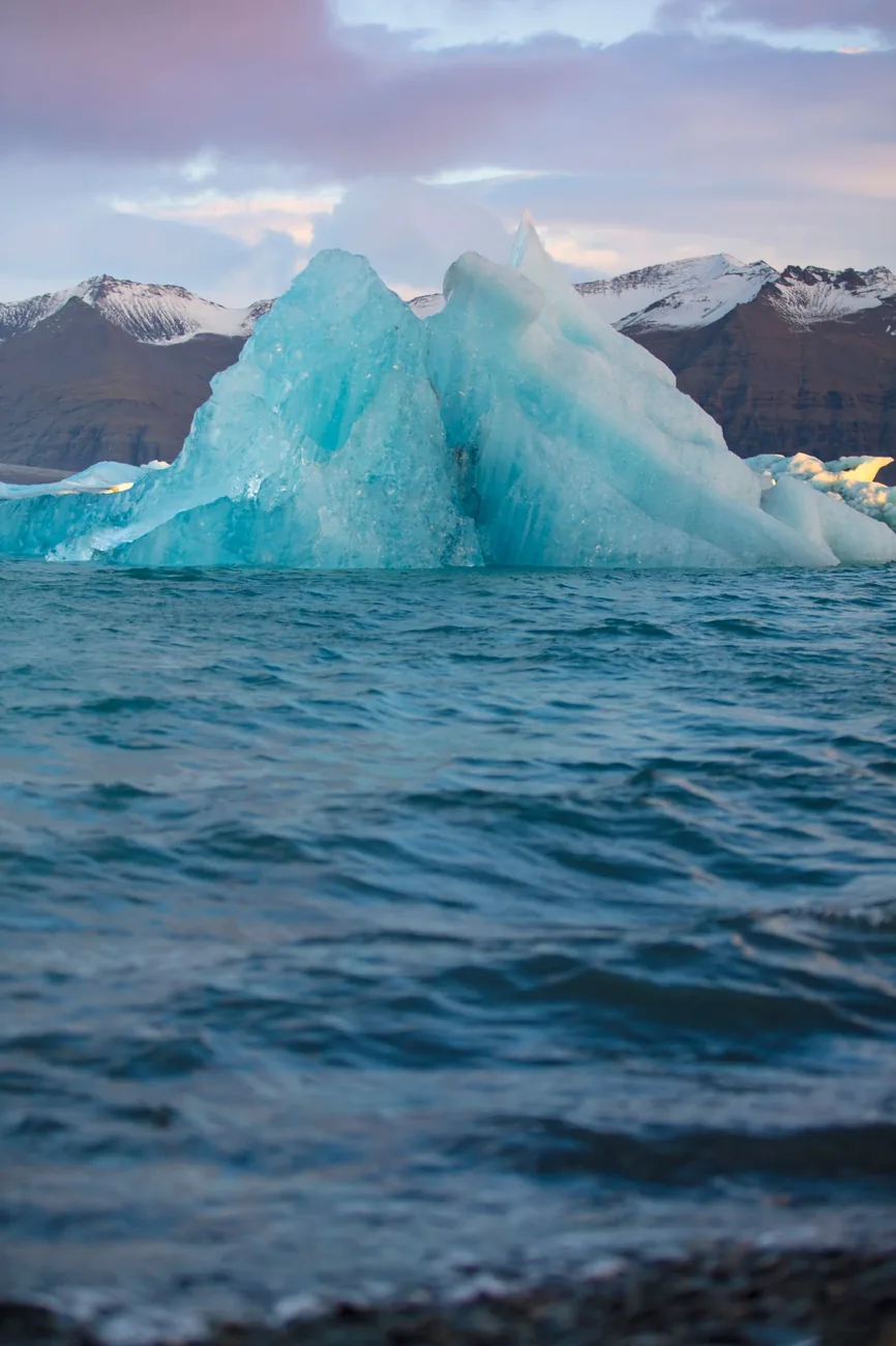 Iceberg azulado flotando en aguas oscuras al anochecer; gran parte del hielo queda bajo la superficie
