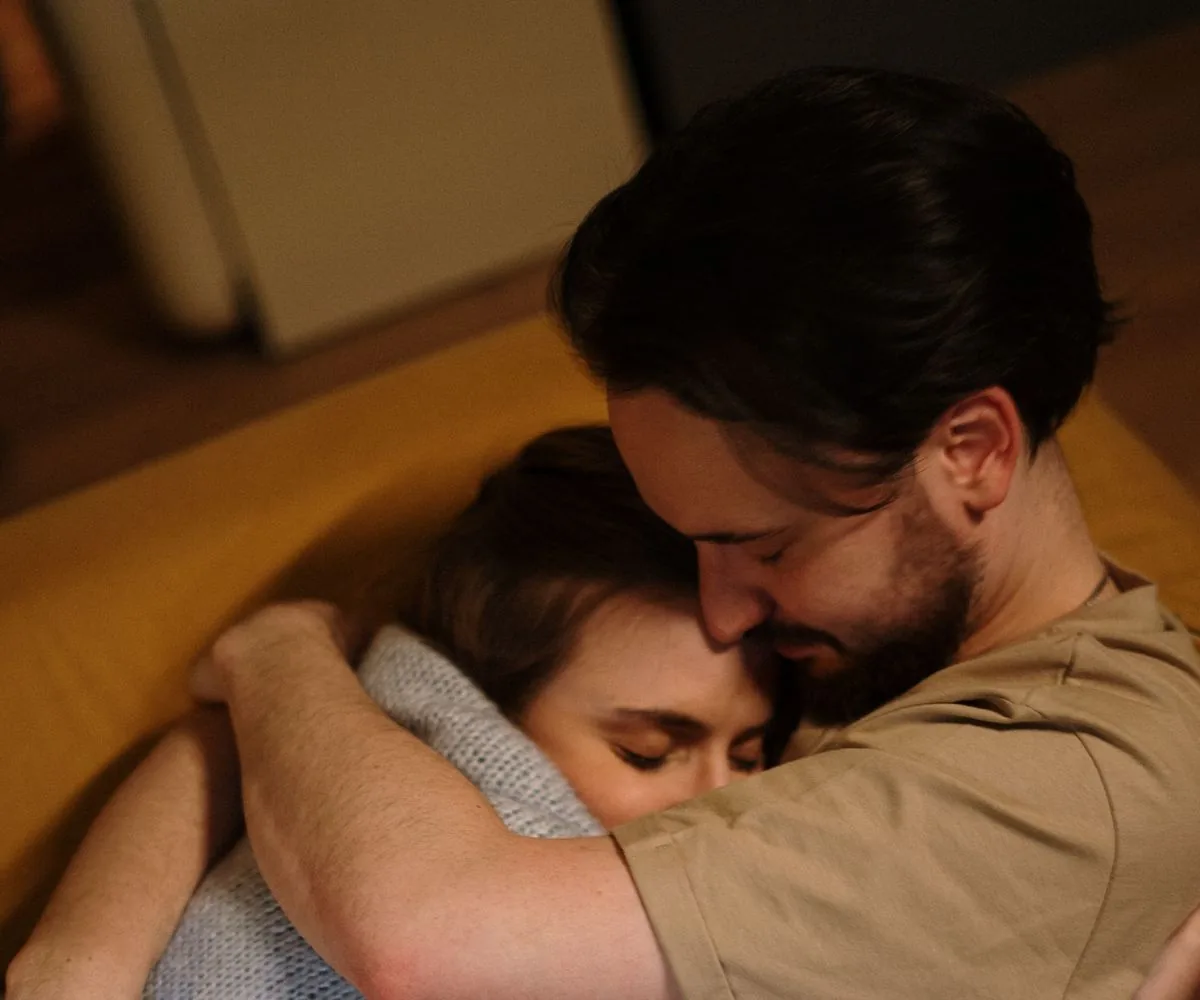 Pareja abrazada en un sofá: él la rodea con los brazos y apoya la cara en su cabeza; los dos con los ojos cerrados