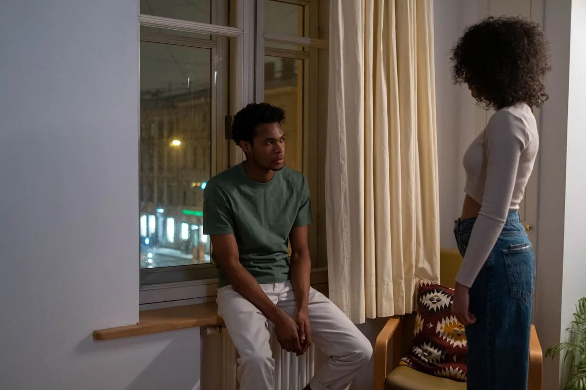 Él sentado junto a la ventana y ella de pie, dándole la espalda, en una habitación con luz cálida