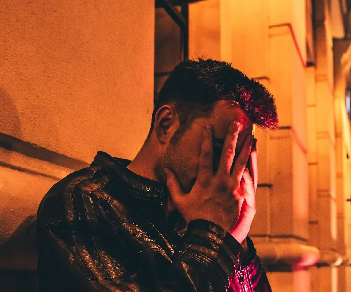 Hombre de noche junto a una pared iluminada, cubriéndose el rostro con la mano