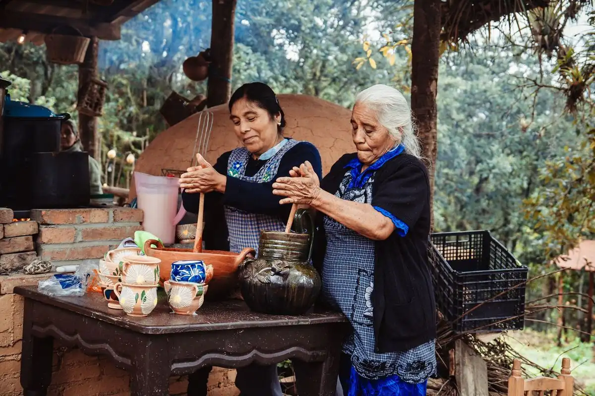 Dos mujeres de distintas generaciones cocinan juntas en una cocina tradicional de leña, con ollas de barro
